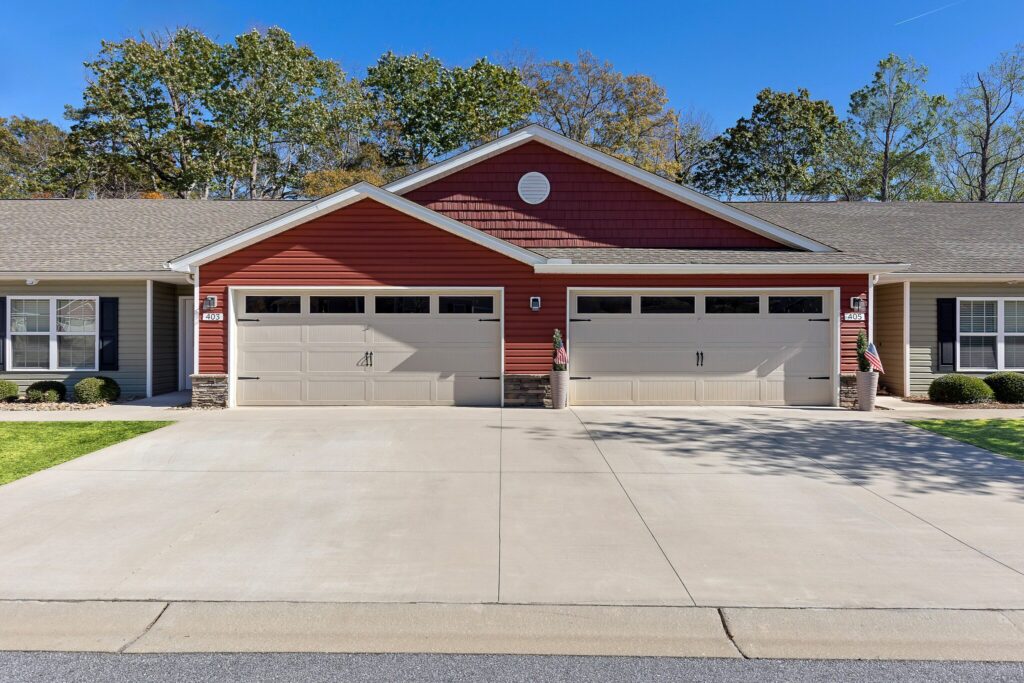 A duplex building with red siding and two double garages, concrete driveway, and manicured lawns, set against a backdrop of trees and blue sky.