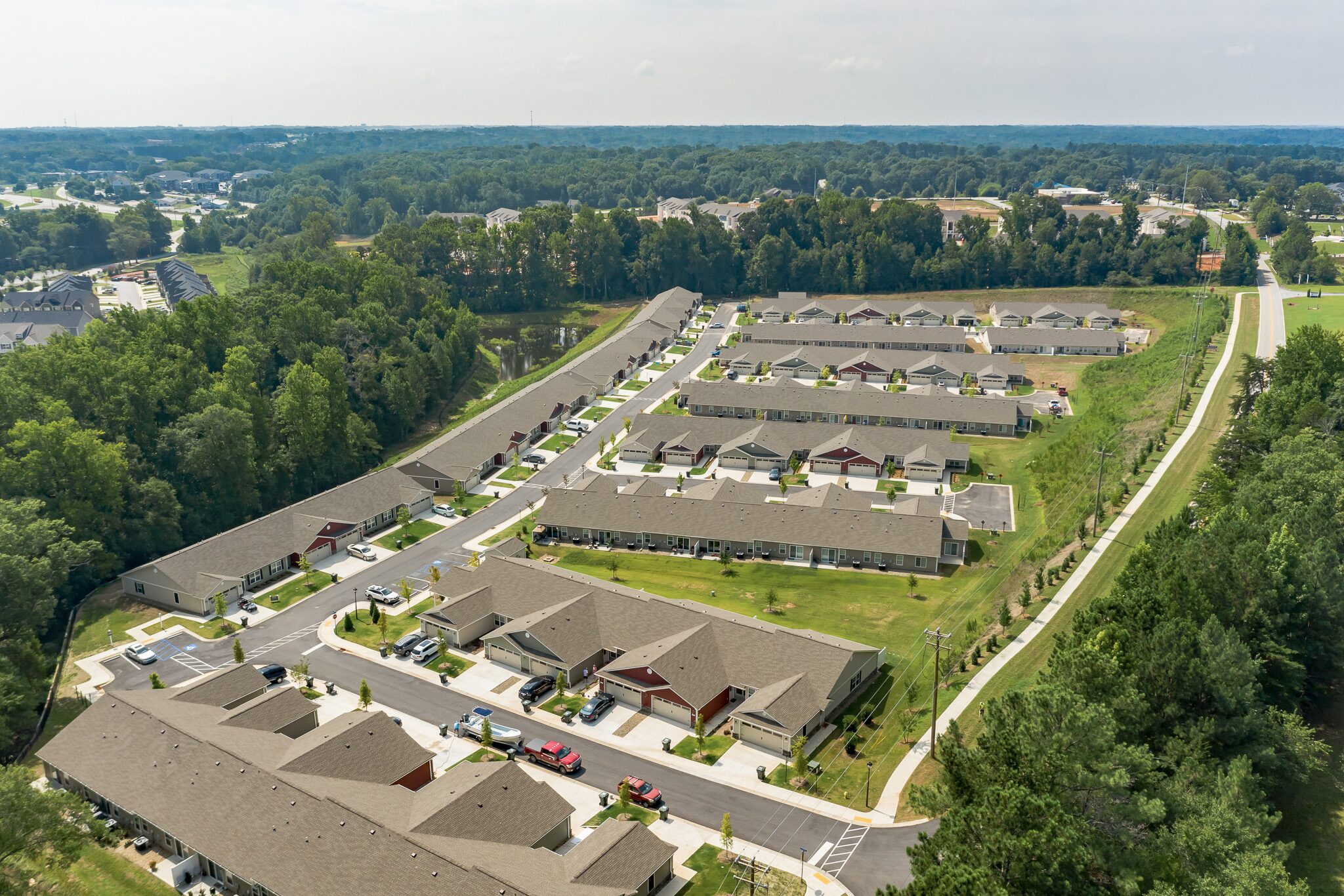 Aerial view of a suburban residential neighborhood with rows of single-story houses, driveways, parked cars, green lawns, and surrounding trees.