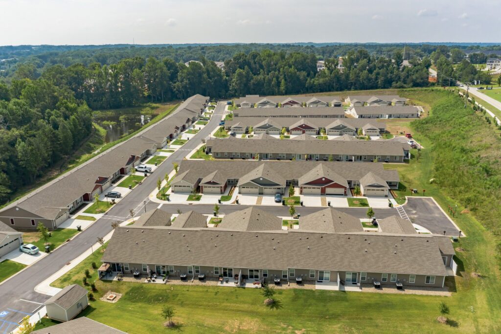 Aerial view of a suburban residential neighborhood with rows of single-story townhouses, driveways, lawns, and surrounding trees.