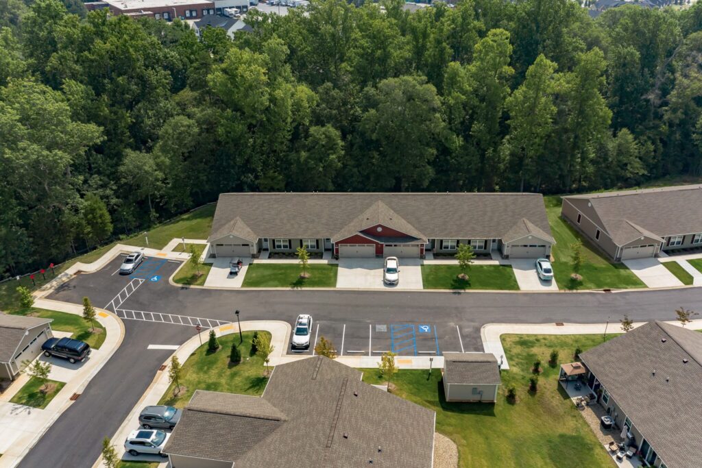 Aerial view of a residential complex with single-story buildings, parked cars, green lawns, sidewalks, and a wooded area in the background.