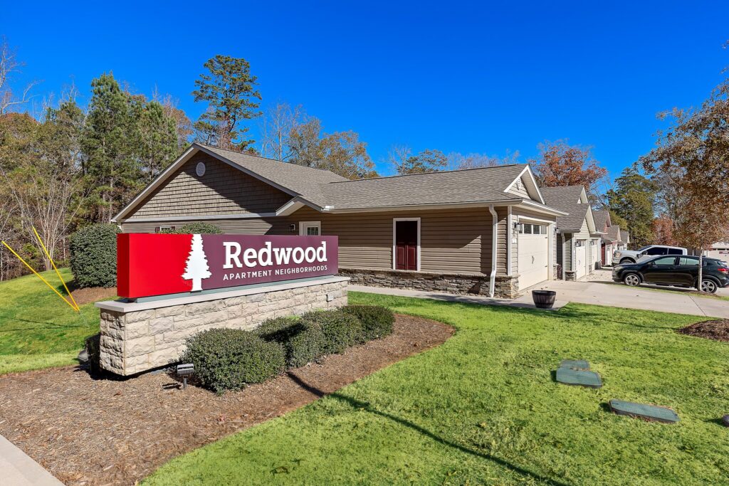 A building with a "Redwood Apartment Neighborhoods" sign in front, surrounded by green grass, shrubs, and parked cars on a sunny day.