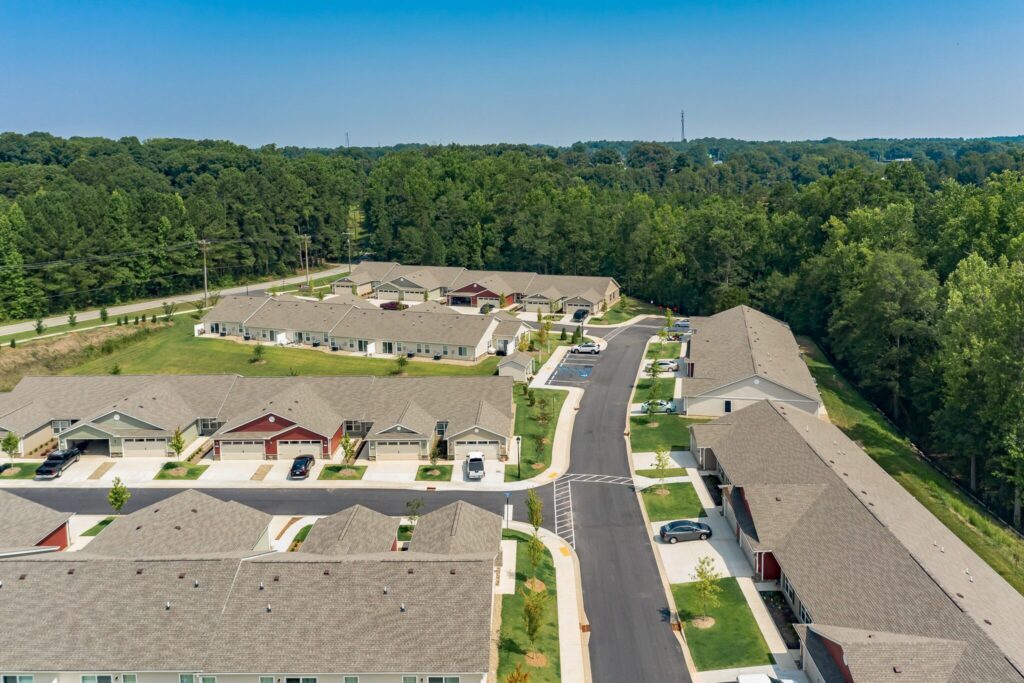 Aerial view of a suburban residential neighborhood with single-story houses, driveways, green lawns, and surrounding trees under a clear sky.