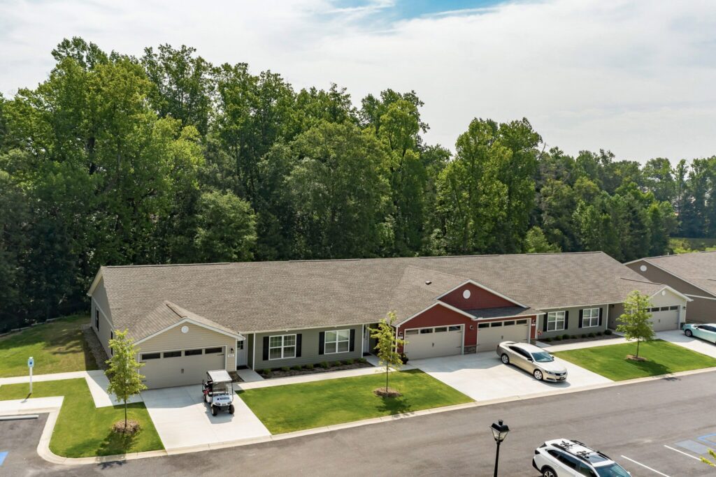 Single-story residential building with attached garages, multiple cars parked in front, and a backdrop of tall trees and greenery.
