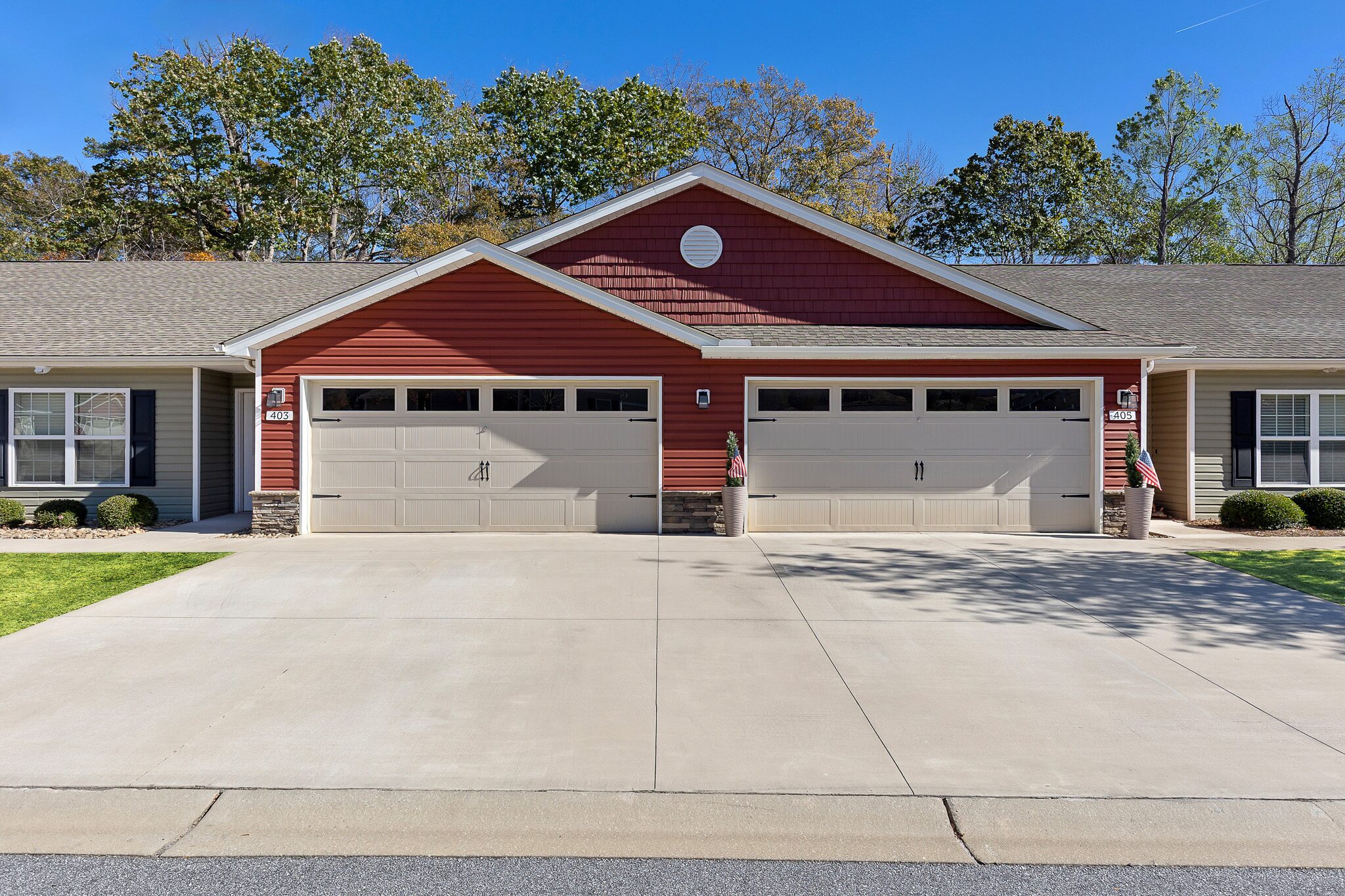 A duplex building with red siding and two double garages, concrete driveway, and manicured lawns, set against a backdrop of trees and blue sky.