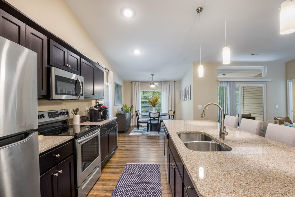 Modern kitchen with dark cabinets, stainless steel appliances, granite countertops, and a breakfast bar, opening to a dining area with large windows and a view of the patio.