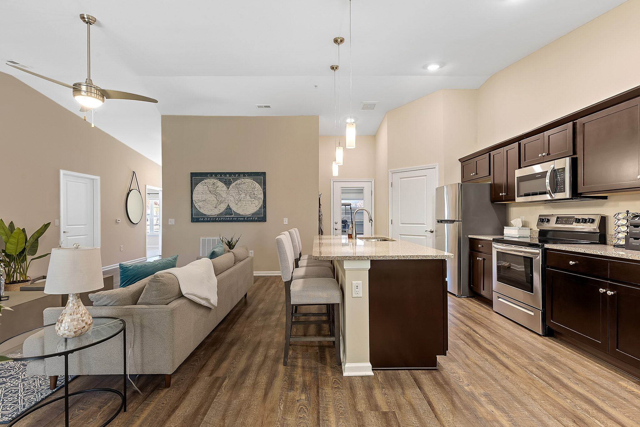 Open-concept kitchen and living room with dark cabinets, stainless steel appliances, granite island, wood flooring, and neutral-colored furniture.