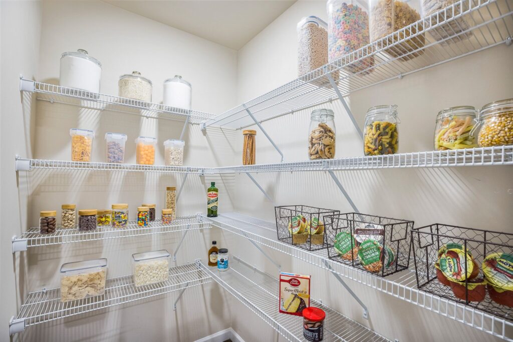 A tidy pantry with wire shelves holding jars of dry goods, canned items, baskets with snacks, and assorted food containers against cream-colored walls.