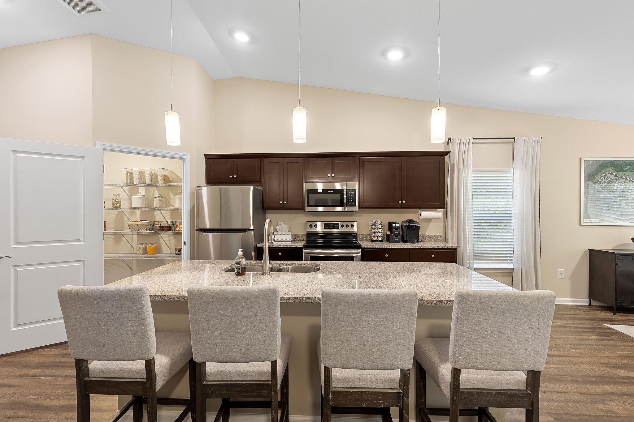 Modern kitchen with a center island, four upholstered chairs, stainless steel appliances, dark wood cabinets, and an open pantry door.