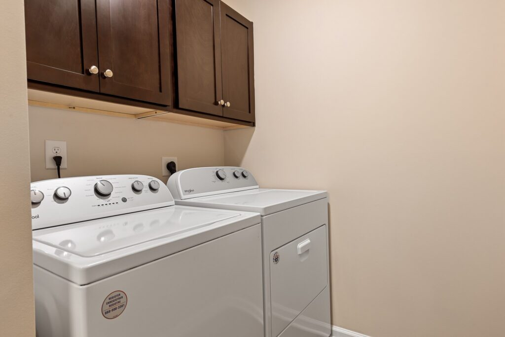 A laundry room with a washing machine and dryer side by side beneath wooden cabinets against a beige wall.