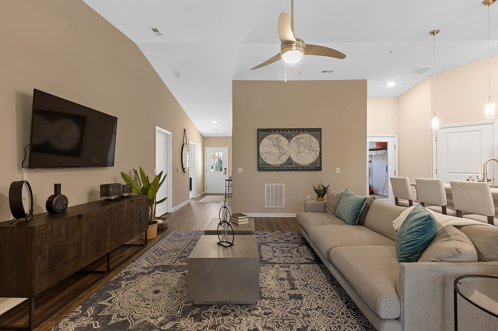 Modern living room with beige walls, a large gray sectional sofa, blue patterned rug, wall-mounted TV, and contemporary decor. Kitchen and dining area visible in the background.