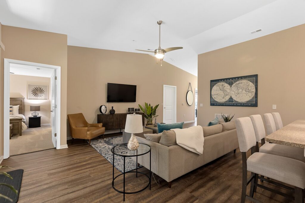 Modern living room with beige walls, a gray sectional sofa, armchair, TV mounted on the wall, bar stools at a kitchen counter, and a map artwork above the sofa.