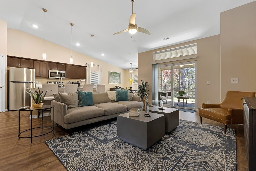 Modern open-plan living room and kitchen with neutral tones, a gray sofa, armchair, pendant lights, patterned rug, and large windows providing natural light and outdoor view.
