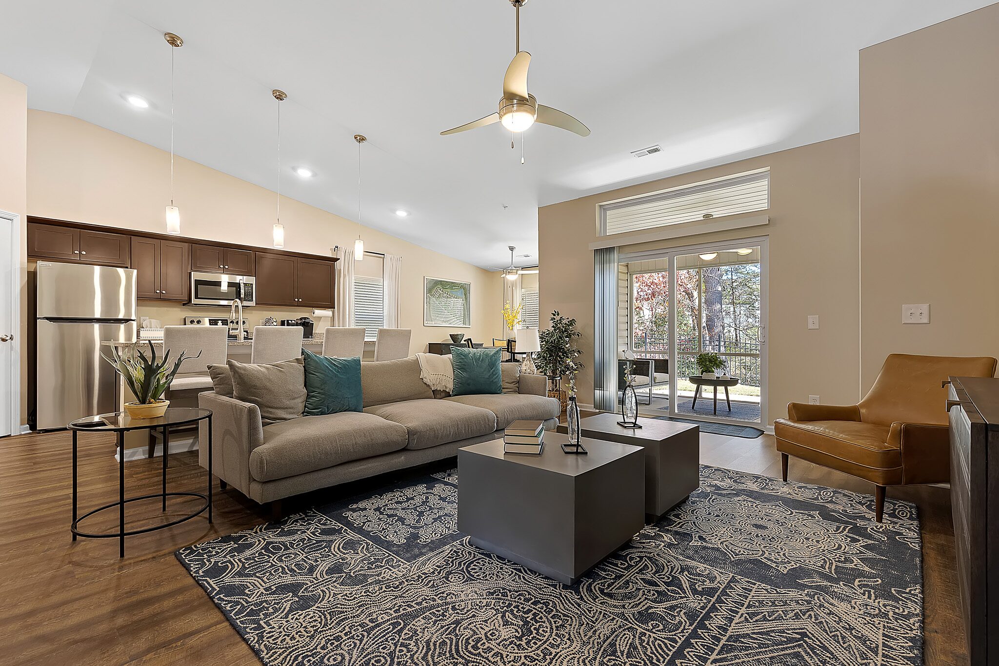 Modern open-plan living room and kitchen with neutral tones, a gray sofa, armchair, pendant lights, patterned rug, and large windows providing natural light and outdoor view.