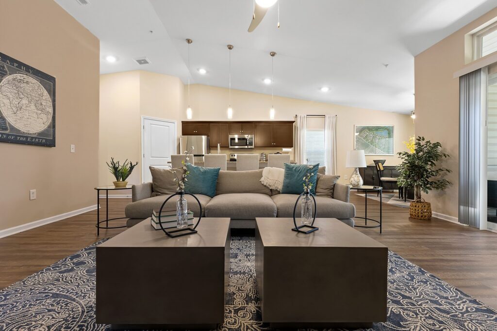 Modern living room with a gray sofa, blue accent pillows, two square coffee tables, and an open view of a kitchen and dining area in the background.
