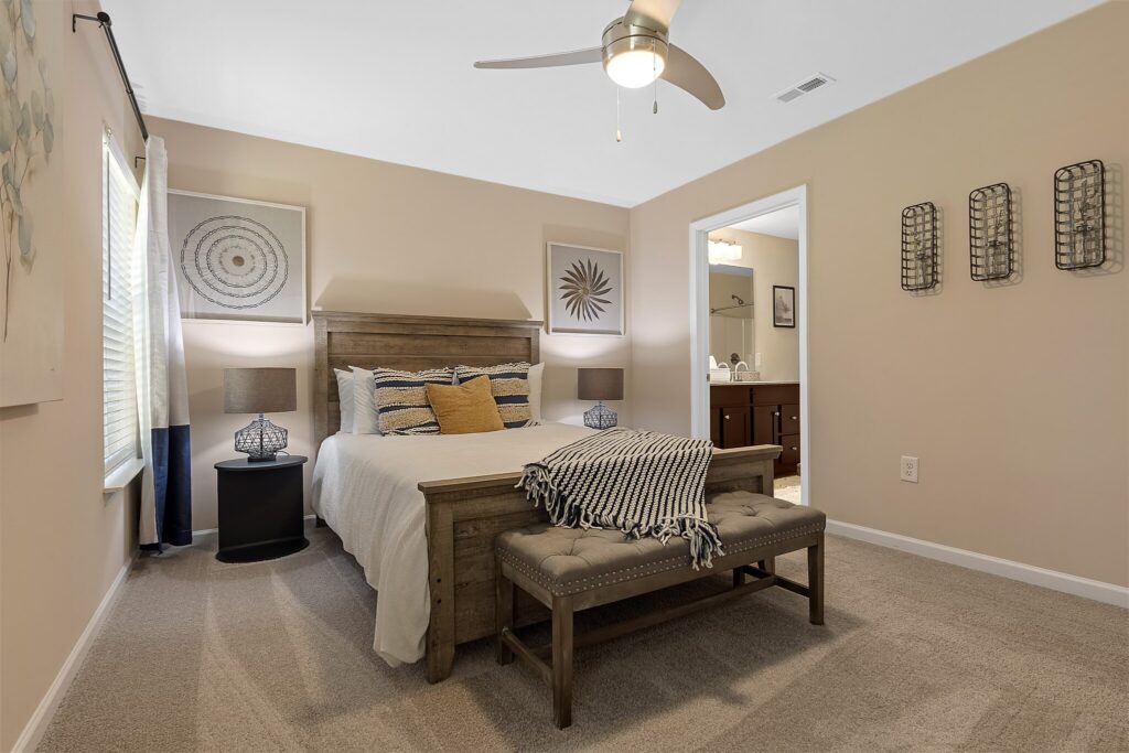 A neatly arranged bedroom with a wooden bed, beige walls, carpeted floor, bench at the foot of the bed, two nightstands, and a doorway leading to a bathroom.
