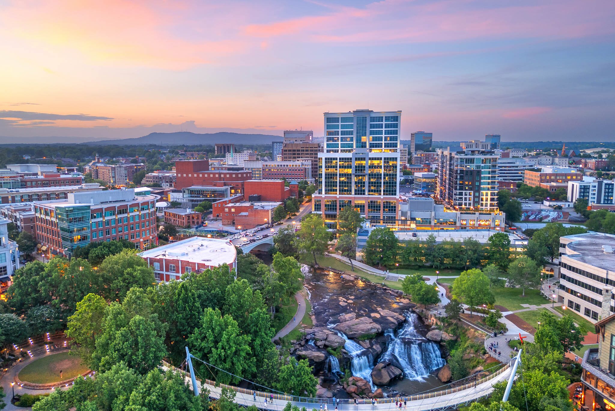 Aerial view of a cityscape at sunset featuring modern buildings, a river with waterfalls, a pedestrian bridge, and green spaces.