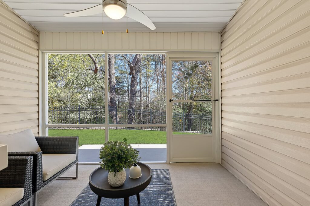 Screened-in porch with beige siding, a ceiling fan, outdoor furniture, a small table with plants, and a view of a grassy yard and trees through large windows.