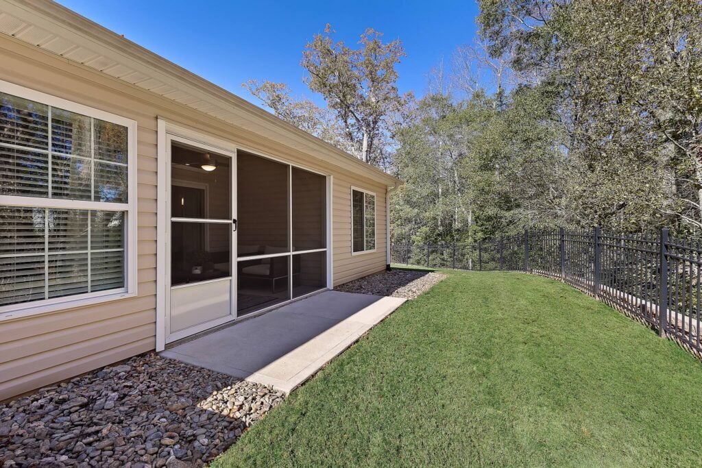 A house with beige siding features a sliding glass door opening to a small concrete patio, a narrow lawn, and a black metal fence bordered by trees.