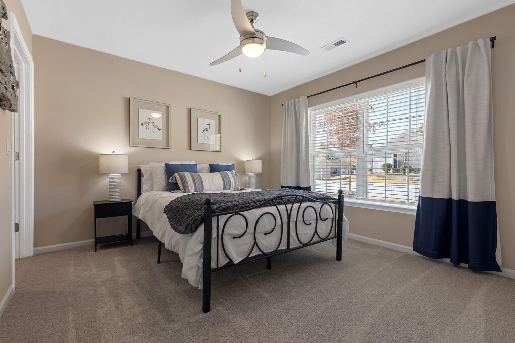 Bedroom with a black metal bed frame, gray bedding, two nightstands with lamps, framed art on the wall, beige carpet, and a large window with white curtains.