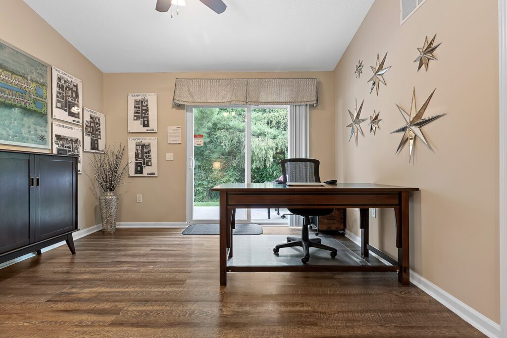 A home office with a wooden desk, office chair, wall art featuring stars, framed floor plans on the walls, a cabinet, and sliding glass doors opening to a green outdoor area.