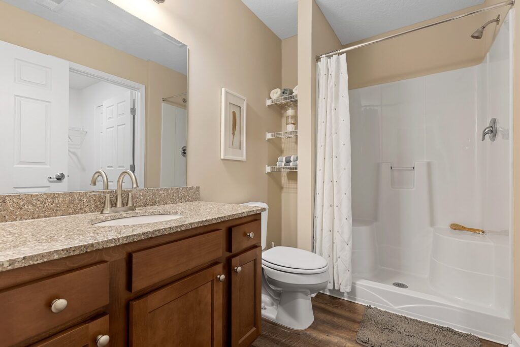 A bathroom with a granite countertop, wooden vanity, toilet, and white shower stall with a curtain, decorated with shelves and neutral wall art.