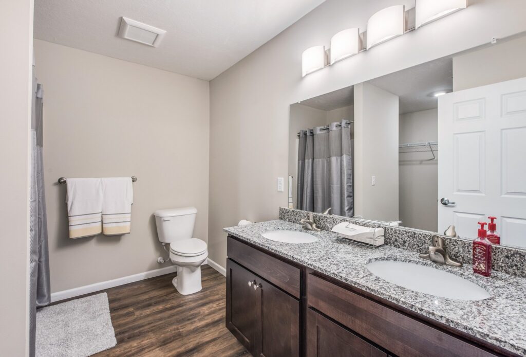 Modern bathroom with double sink vanity, granite countertop, dark wood cabinets, toilet, towel rack, shower with gray curtain, and wood-style flooring.