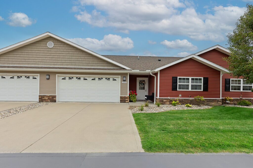 Single-story house with a red and beige exterior, attached three-car garage, concrete driveway, and landscaped front yard under a partly cloudy sky.
