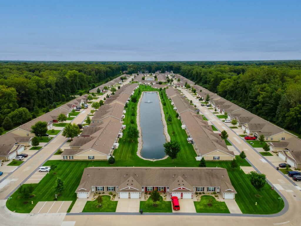 Aerial view of a suburban neighborhood with rows of houses and a long central pond, surrounded by green lawns and bordered by trees.