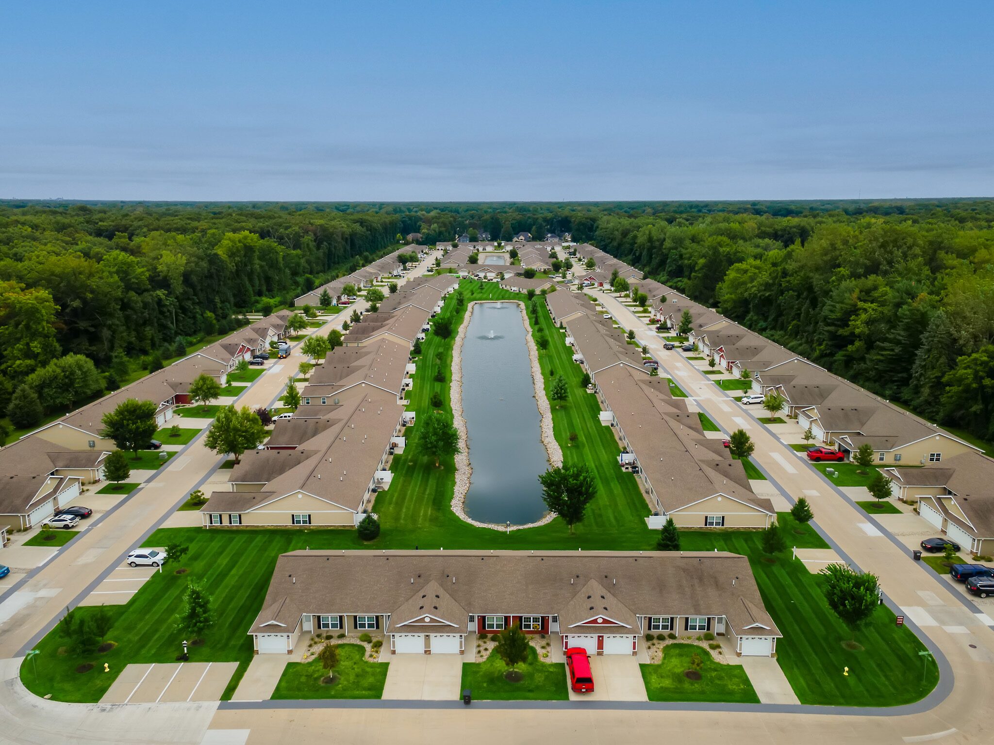 Aerial view of a suburban neighborhood with rows of houses and a long central pond, surrounded by green lawns and bordered by trees.