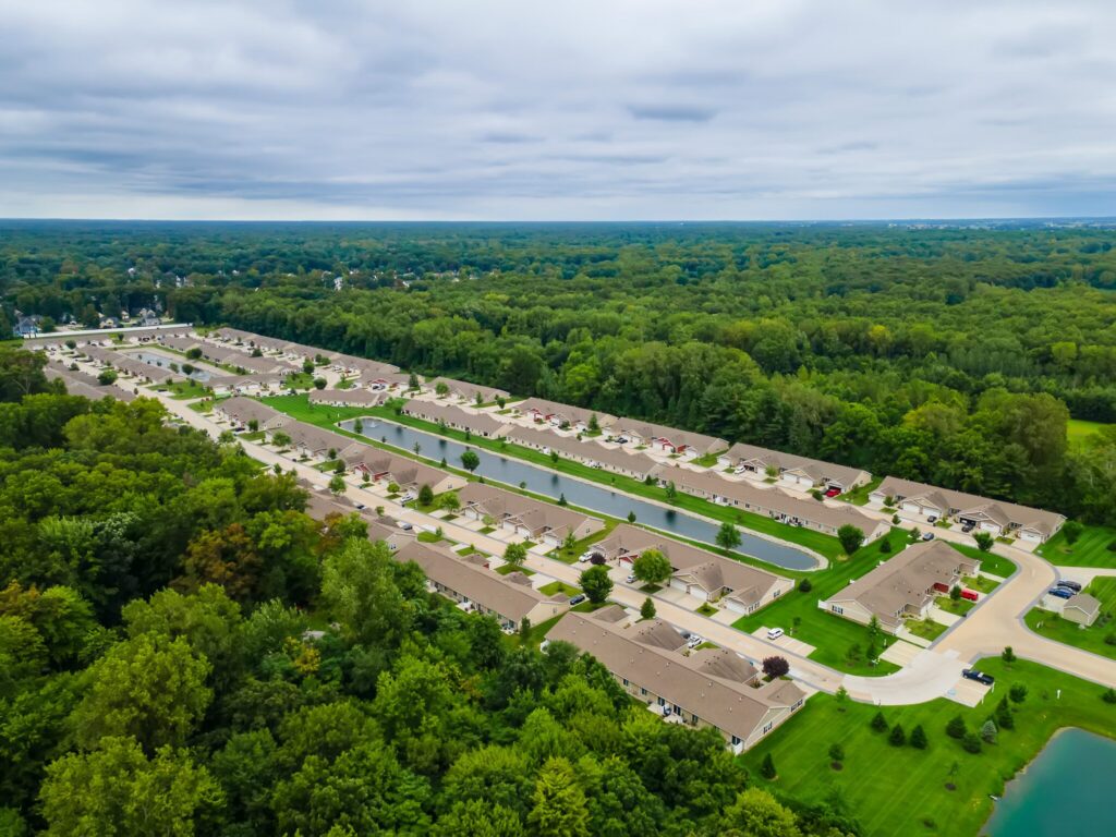 Aerial view of a suburban neighborhood with rows of houses, green lawns, streets, a pond, and dense forest in the background under a cloudy sky.