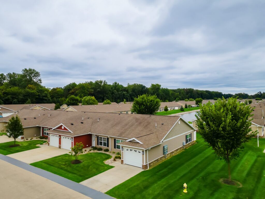 A row of single-story suburban houses with garages, green lawns, and trees under a cloudy sky.