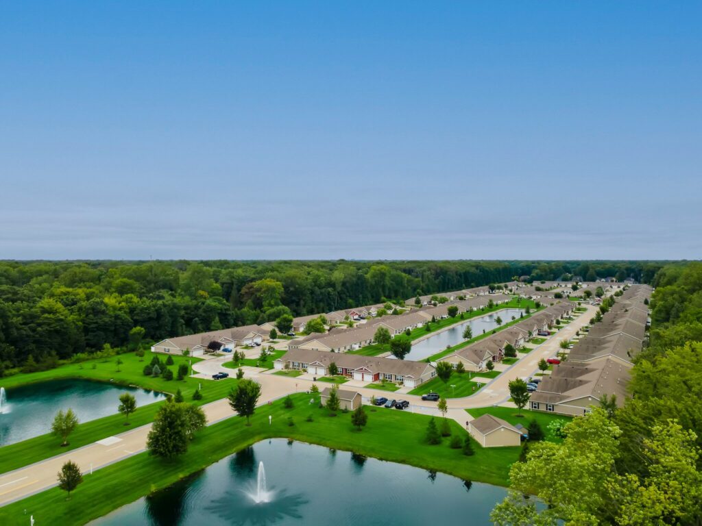 Aerial view of a suburban neighborhood with rows of houses, small ponds, green lawns, and a fountain near a wooded area under a clear blue sky.