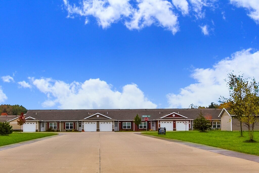 Single-story residential buildings with attached garages line both sides of a wide driveway under a bright blue sky with scattered clouds.