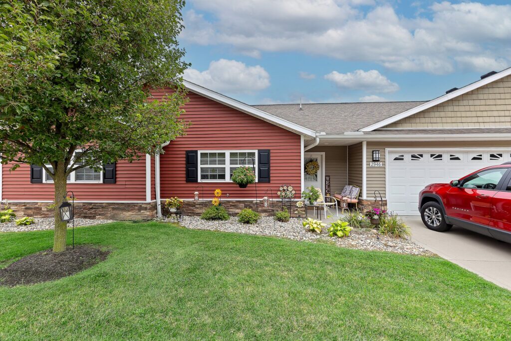 Single-story house with red siding, black shutters, a manicured lawn, landscaped flower beds, and a red car parked in the driveway under a partly cloudy sky.