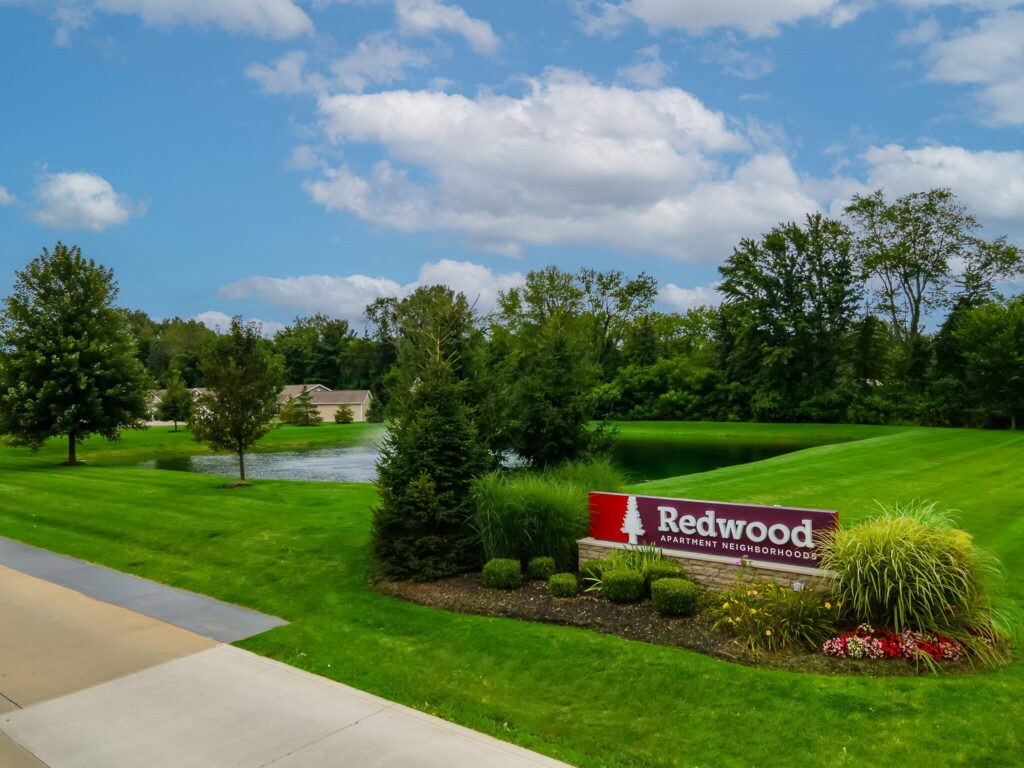 A landscaped entrance with a "Redwood Apartment Neighborhoods" sign, green lawn, pond, trees, and partly cloudy sky.