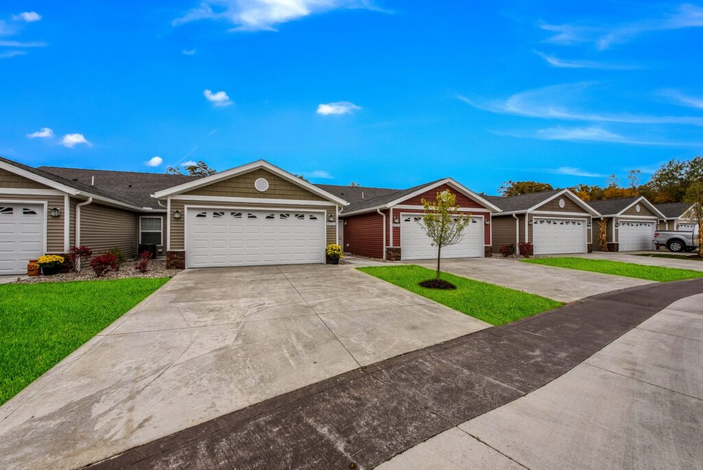 Row of modern single-story houses with attached garages, concrete driveways, and small front lawns under a bright blue sky.