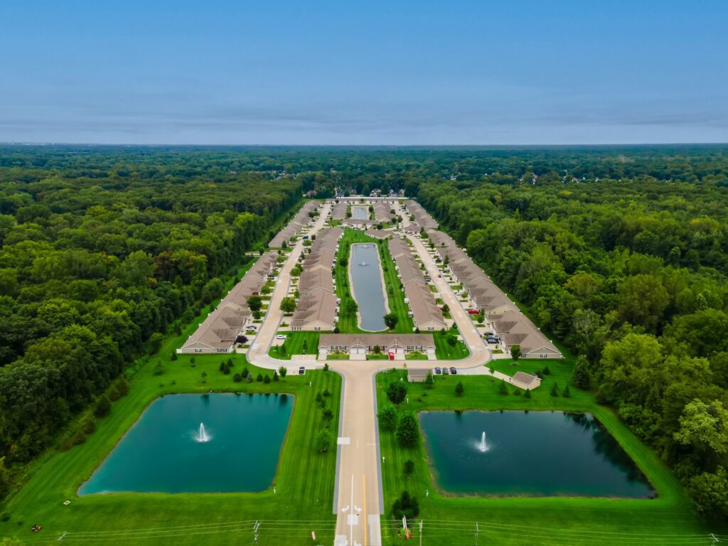 Aerial view of a residential community with multiple houses, two rectangular ponds with fountains at the entrance, and surrounded by dense green trees.
