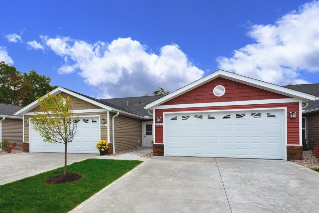 Two suburban houses with white double garages, red and tan siding, and a shared driveway. A small tree and green lawn are in front of the homes under a partly cloudy sky.