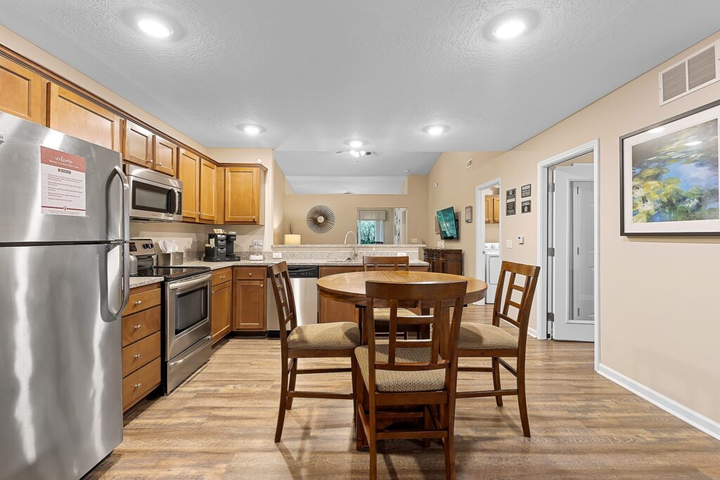 Modern kitchen and dining area with stainless steel appliances, wooden cabinets, round dining table with four chairs, and hardwood flooring.