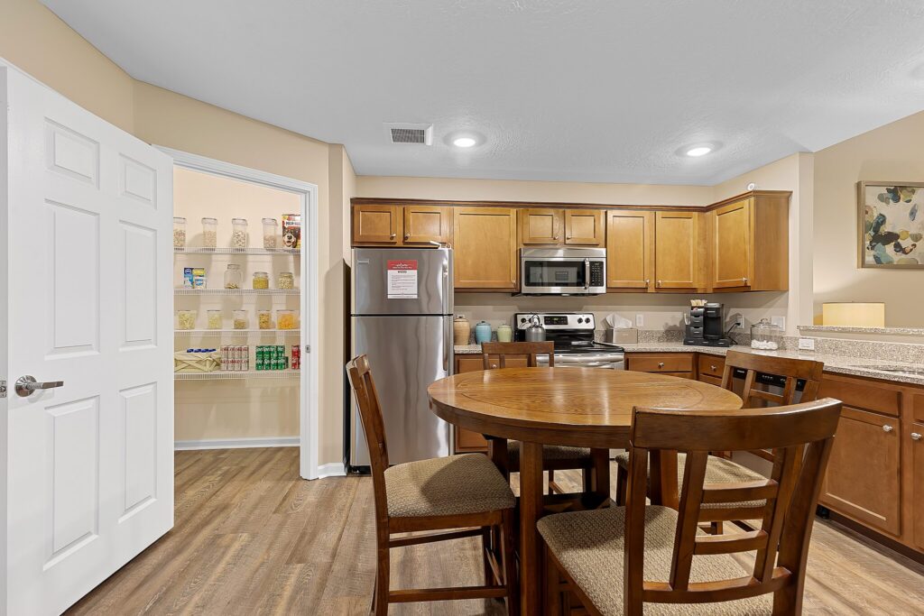 Modern kitchen with wooden cabinets, stainless steel appliances, a round dining table with four chairs, and an open pantry stocked with containers and food items.