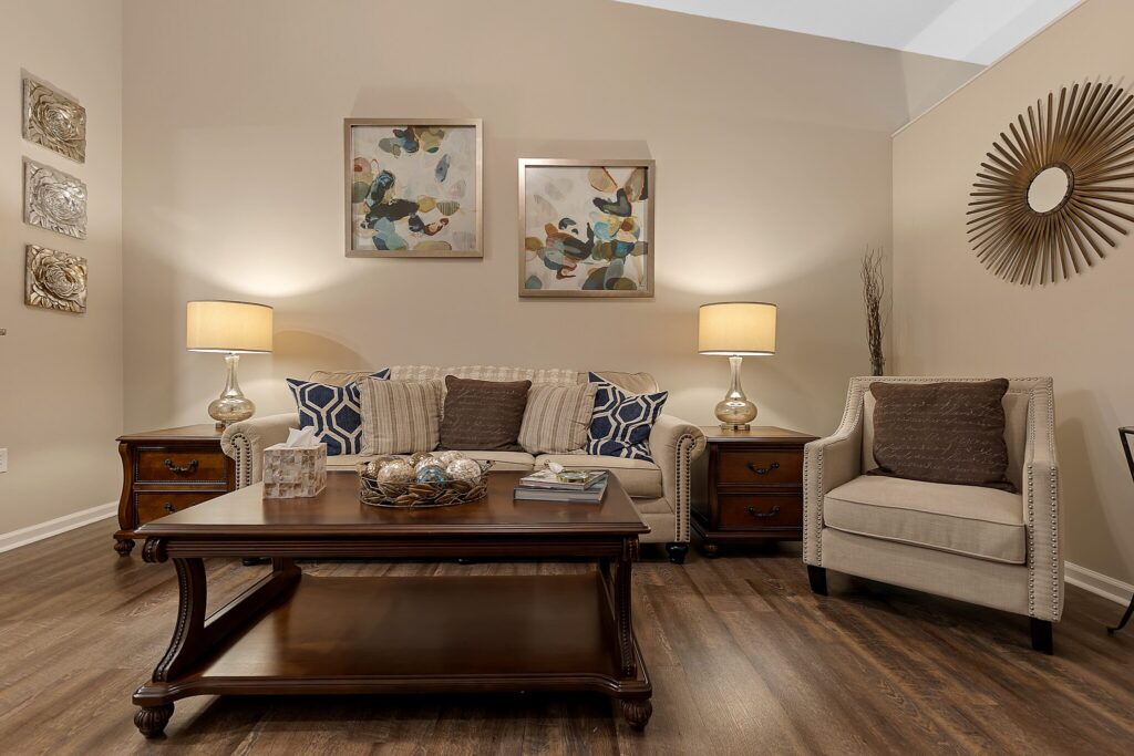 A living room with a beige sofa, armchair, wooden coffee table, two side tables with lamps, abstract wall art, and a sunburst mirror on hardwood floors.