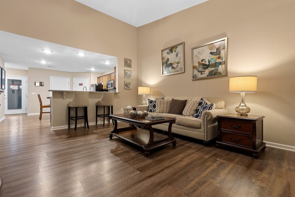 Open-plan living room with a beige sofa, two side tables with lamps, a wooden coffee table, modern artwork, and a kitchen area with a breakfast bar and two chairs in the background.