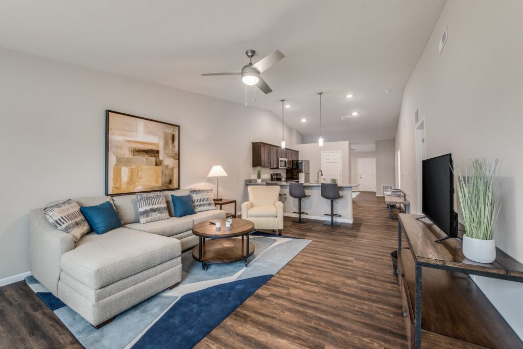 Modern open-plan living room and kitchen with a sectional sofa, TV, bar stools, wall art, and wood flooring under recessed lighting.