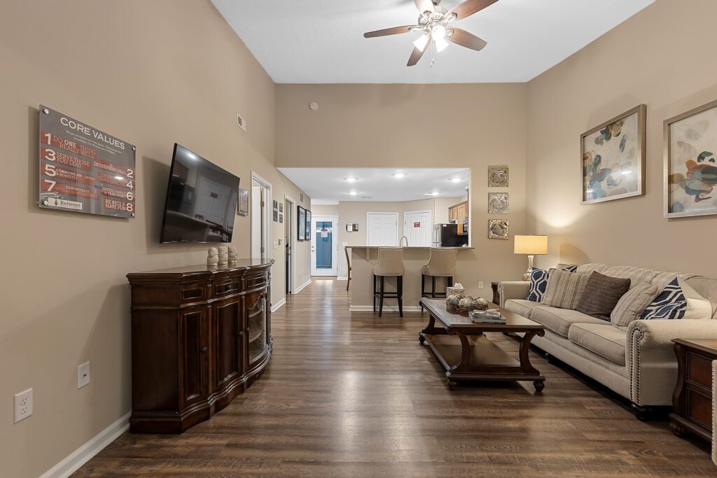A living room with wood floors, a beige sofa, coffee table, TV, and wall art, leading to a kitchen with bar stools and overhead lighting.