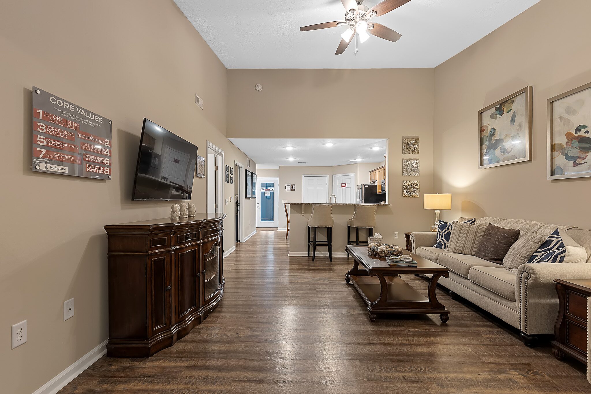 A living room with wood floors, a beige sofa, coffee table, TV, and wall art, leading to a kitchen with bar stools and overhead lighting.