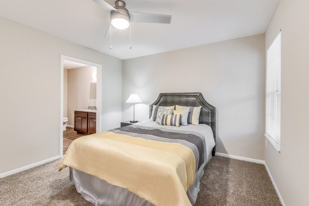 Bedroom with a neatly made bed in striped bedding, a nightstand with a lamp, a ceiling fan, and an open doorway leading to a bathroom.