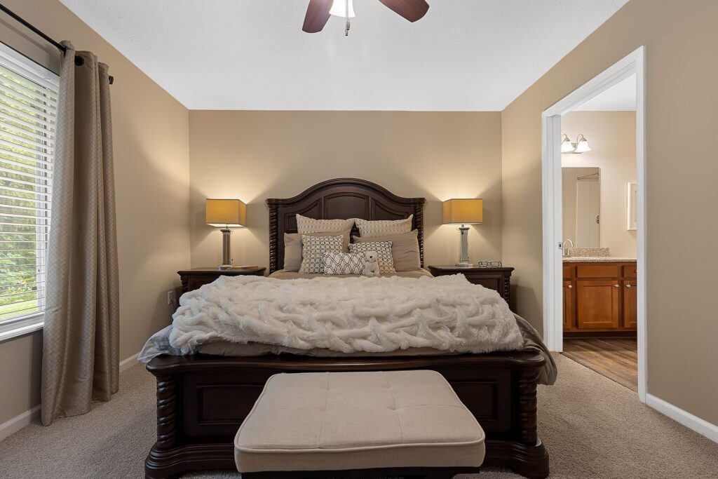A neatly made bed with multiple pillows and a fluffy blanket is centered in a beige bedroom with matching nightstands, lamps, and an open door leading to a bathroom.