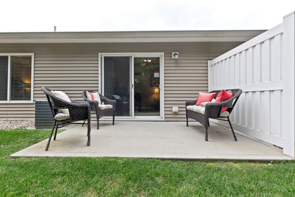 Outdoor patio with two wicker chairs and a loveseat arranged on a concrete slab, bordered by grass and a white privacy fence, in front of a house with sliding glass doors.