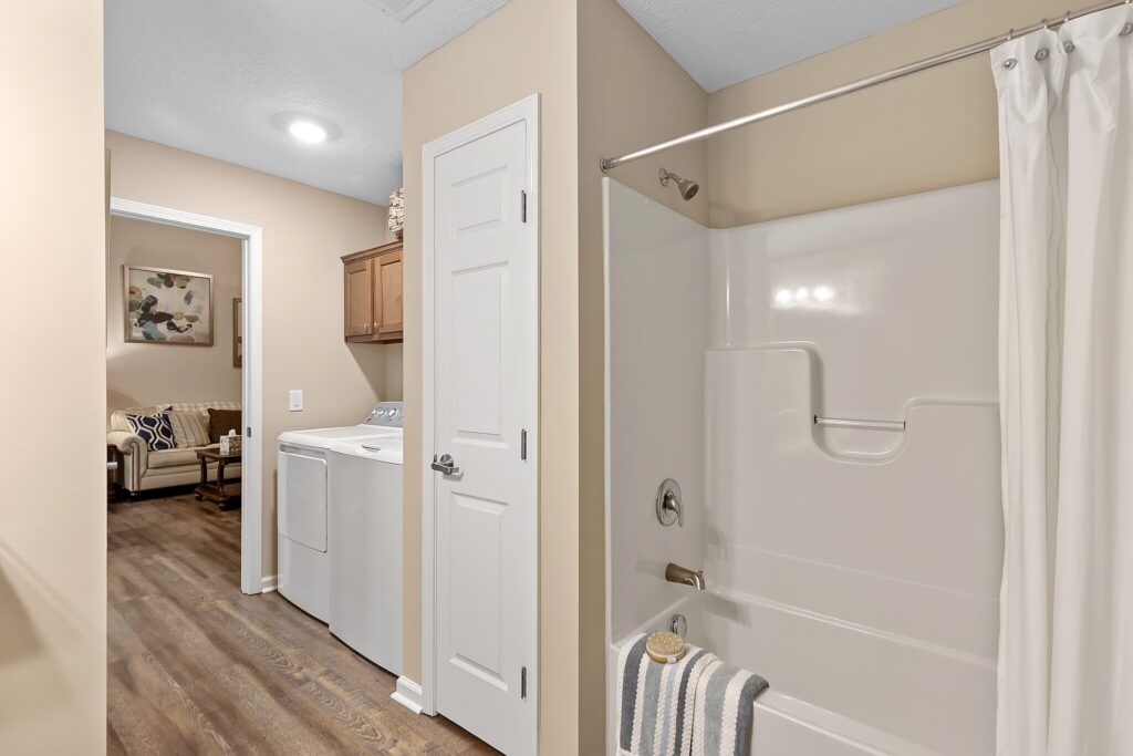 Laundry area with a washer and dryer next to a white door; adjacent is a bathroom with a white bathtub, shower curtain, and a striped towel hanging over the tub edge.