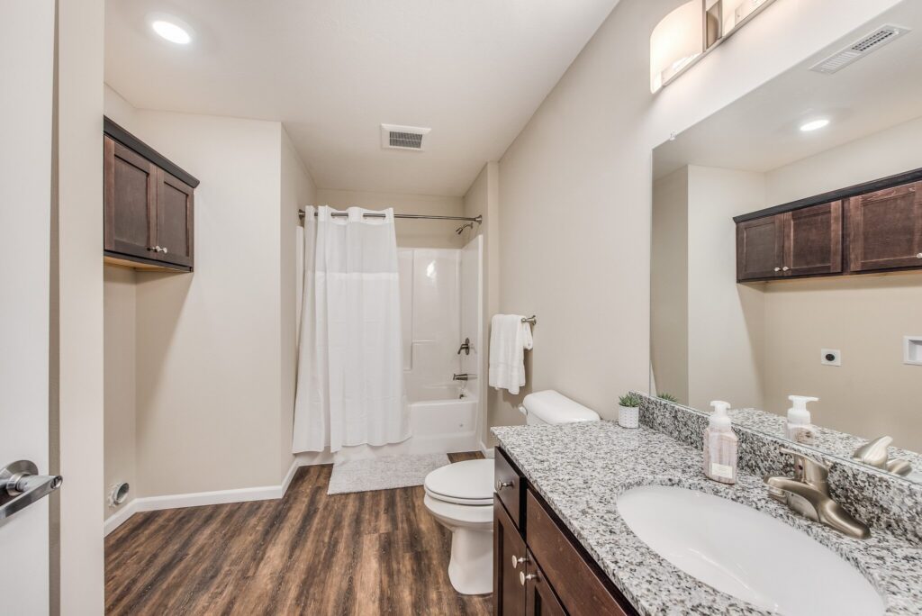 Modern bathroom with wood-look flooring, granite countertop, under-mount sink, toilet, bathtub with white shower curtain, and dark wood cabinets. Bright lighting above the mirror.